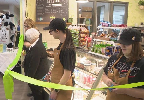 Two young female adults dressed in matching brown aprons and baseball hats cut a green ribbon alongside two Autism Up female staff members, also cutting the green ribbon for the grand opening of PercUp cafe, located inside AutismUp.